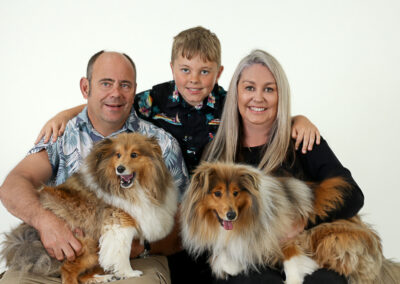 Happy family photo with their pet dogs, taken in a professional portrait studio. Family portrait photography in Auckland featuring mum, dad, son & two collie pet dogs.